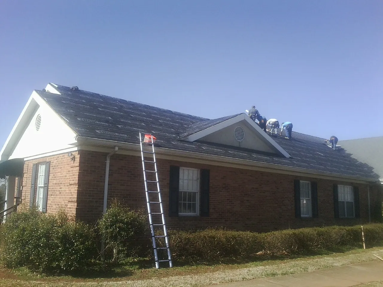 Crew tearing shingles off a brick ranch roof with a ladder set — Metal Roof Repair work by our crew in Erieville