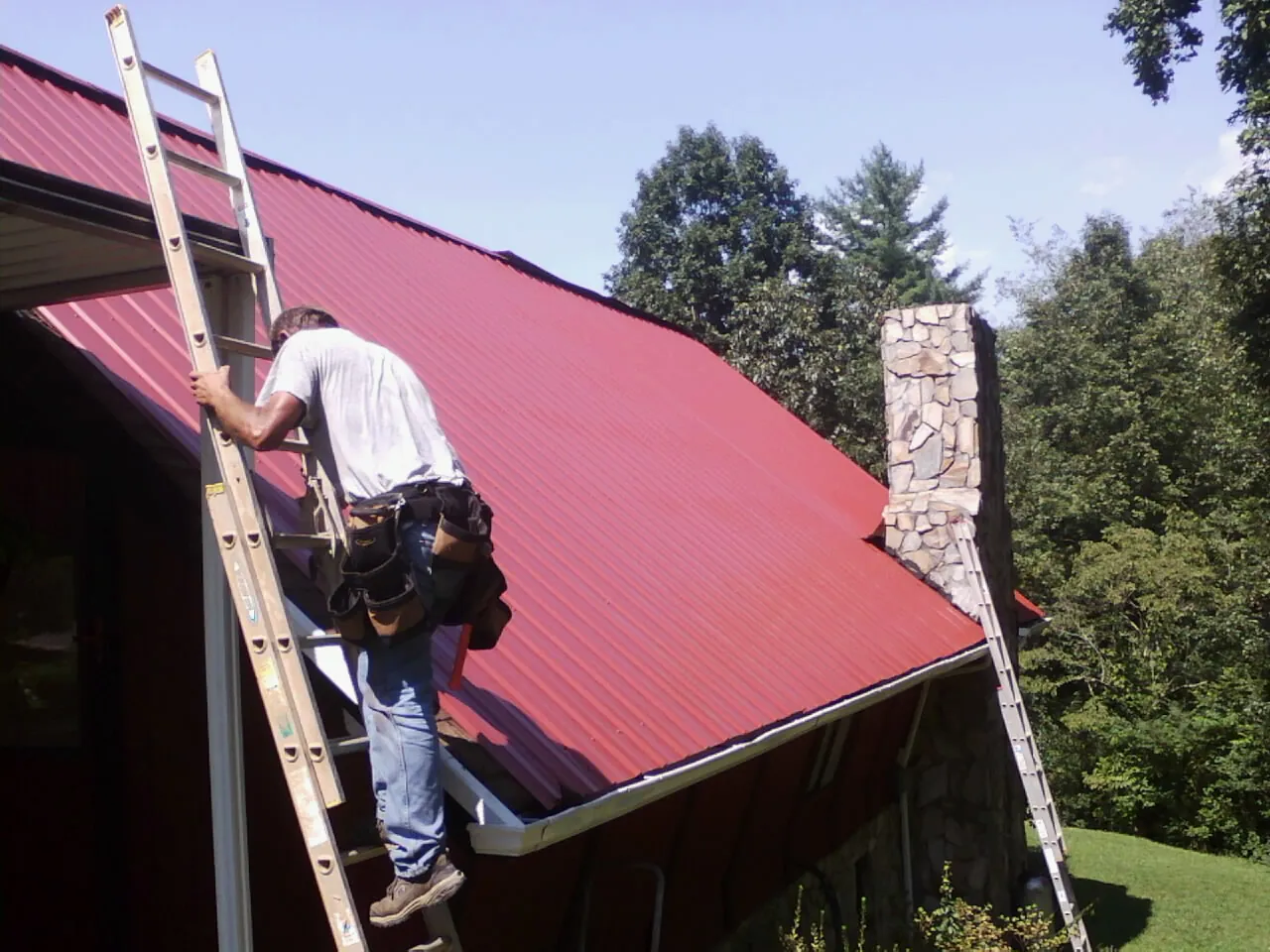 Roofer on a ladder beside a red metal roof and stone chimney — Soffit & Fascia Repair by Roofing Brothers in Erieville